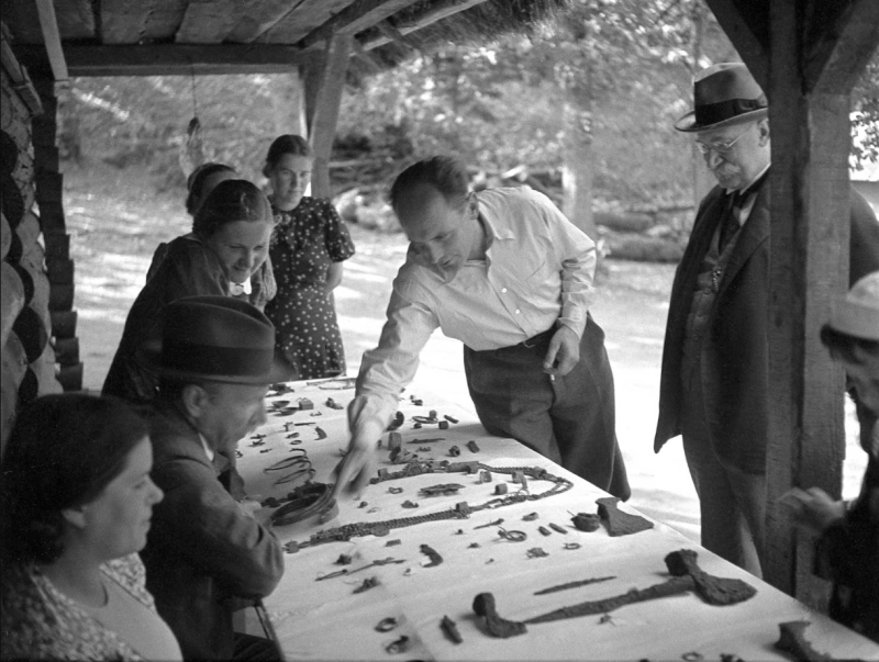 1939. gads. Arheoloģisko izrakumu vadītājs Valdemārs Ģinters (vidū) Mežotnes pilskalnā izrāda profesoriem (no kreisās) Augustam Tentelim un Francim Balodim iegūtās senlietas arheoloģiskās izpētes laikā (fotogrāfs Pēteris Ārends. Avots: Latvijas Nacionālais vēstures muzejs).